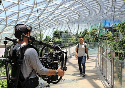 A man wearing jeans, t-shirt, and a backpack in an indoor atrium walks toward another man holding filming equipment. 