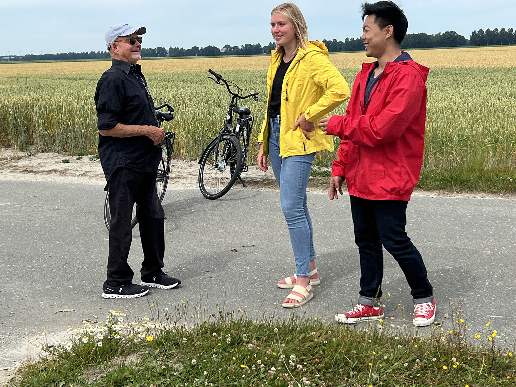 Two men and a woman talk and laugh in a field full of flowers. Two of the people have bikes. 