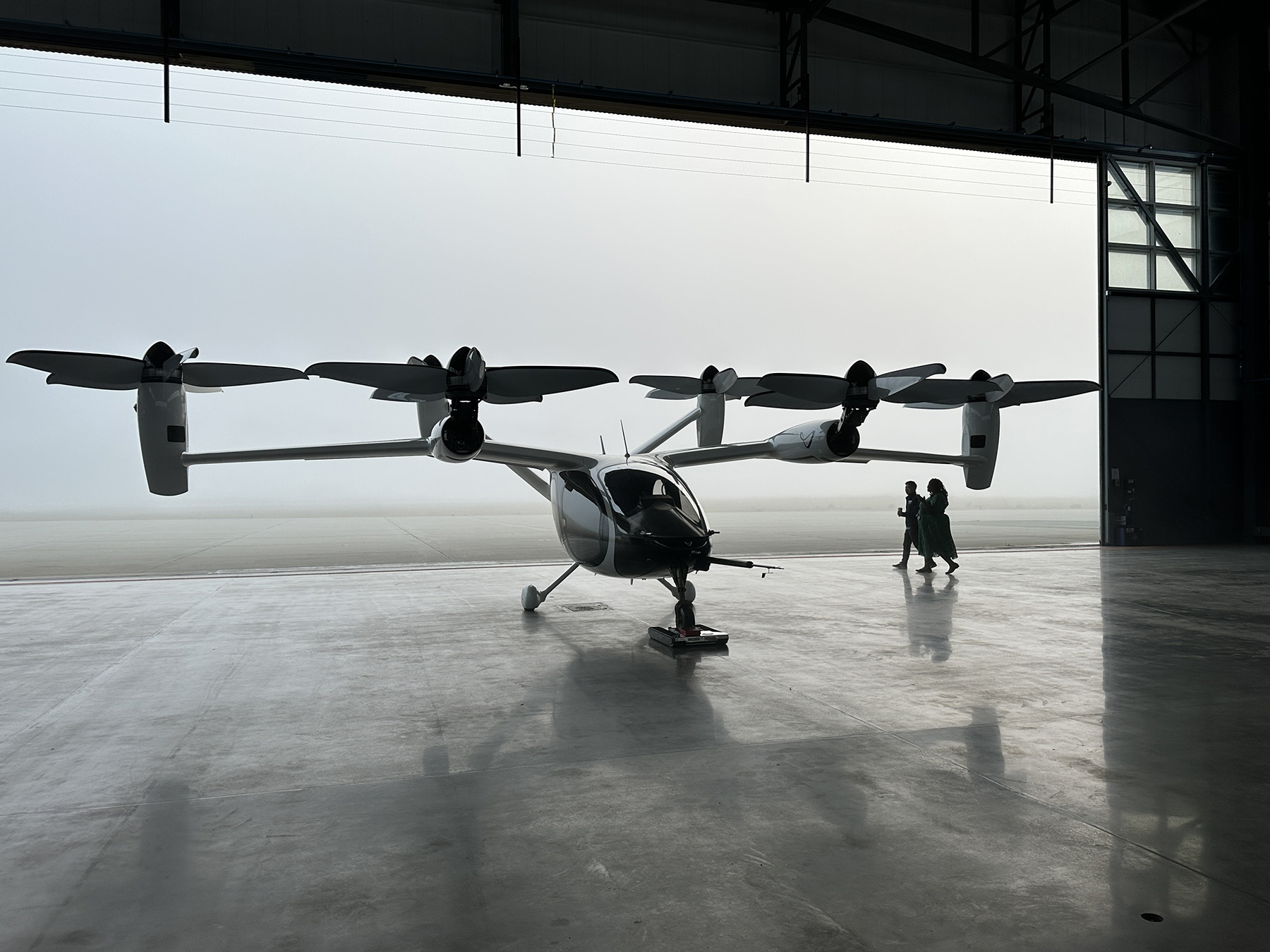 A man and a woman in shadow walk in an aircraft hangar. The aircraft is an electric air taxi that looks like a helicopter but has six motors attached to its wings and other structures. 