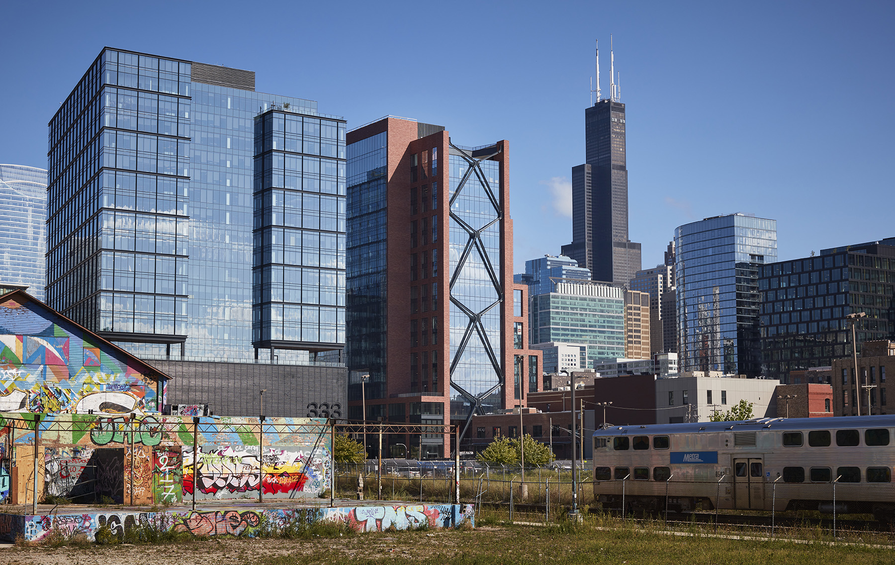 A large multistory, brick, glass, steel, and concrete structure that settles seamlessly with the buildings of various heights that surround it. 