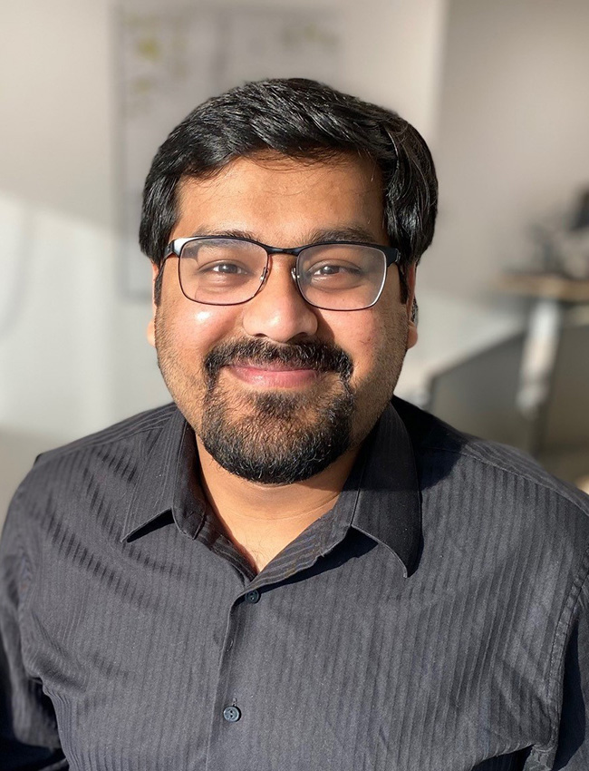 Man with tan skin and dark brown hair wearing a gray shirt and glasses smiles at the camera.