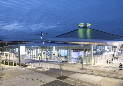 A building with a glass facade has a pointy roof. People walk around outside the building.