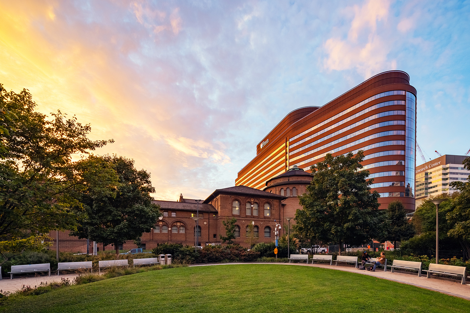 Image shows a multistory building with an oblong shape.  A greenspace with a path and benches is in the foreground.