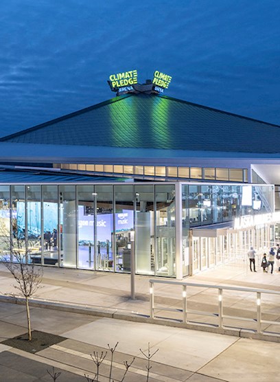 A building with a glass facade has a pointy roof. People walk around outside the building. 