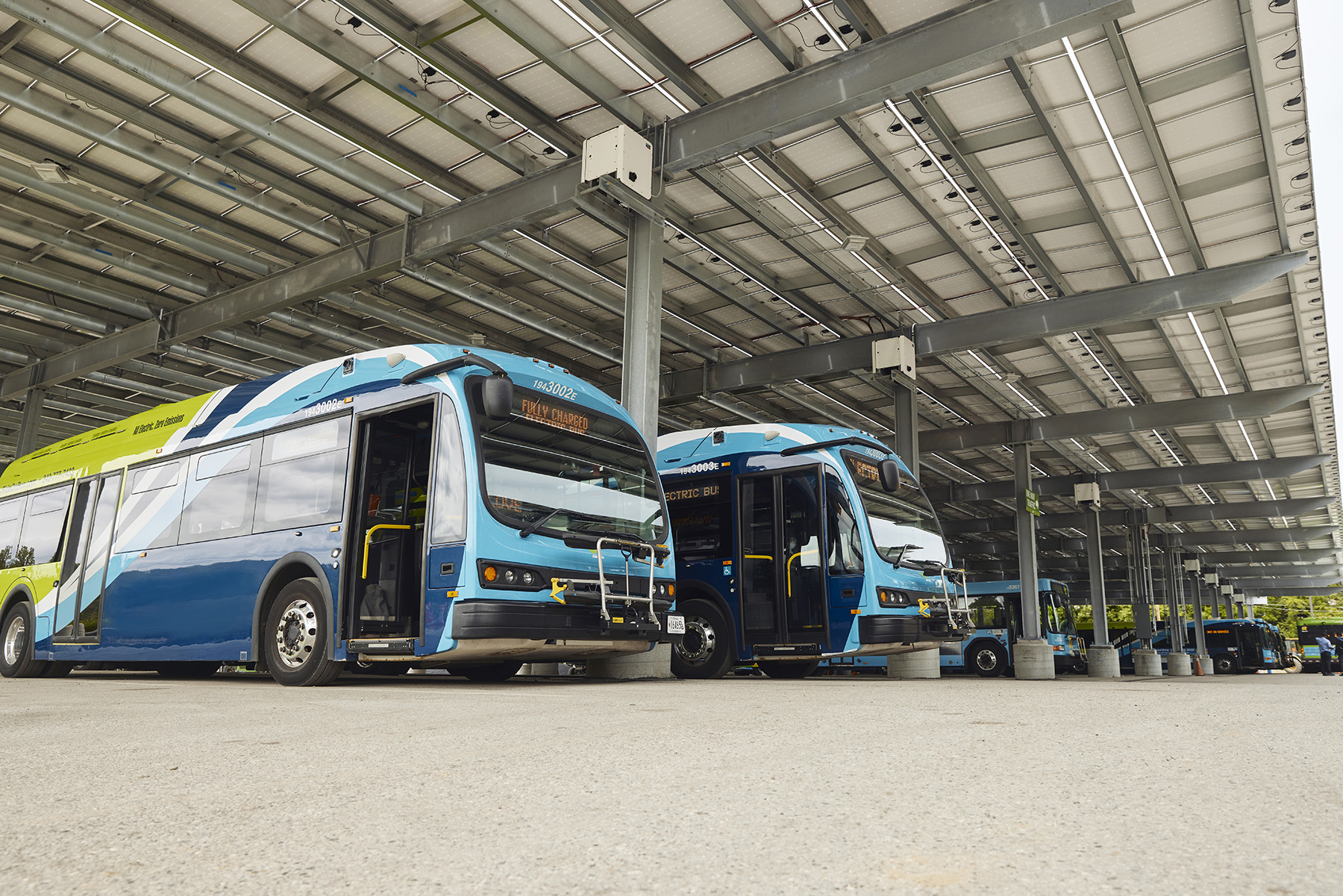 The photograph shows electric buses beneath a canopy structure. 