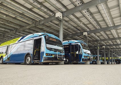 The photograph shows electric buses beneath a canopy structure. 