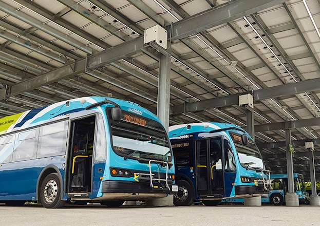 The photograph shows electric buses beneath a canopy structure.