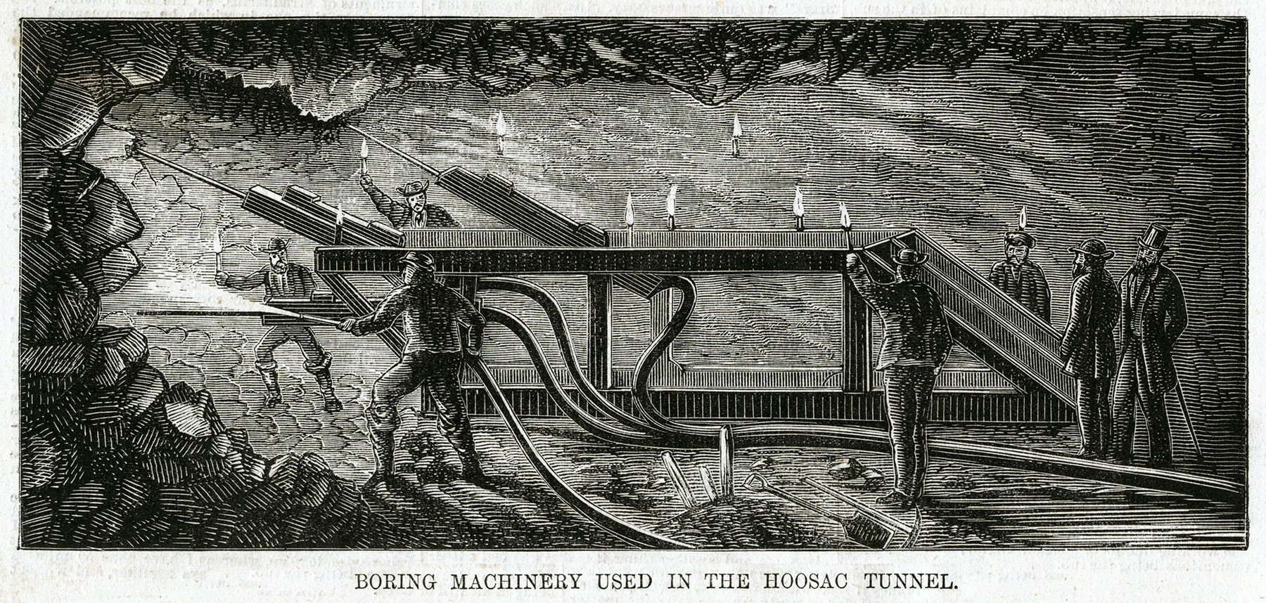 Men hold hoses that are used to blast away rock. Other men hold torches to light the explosive. 