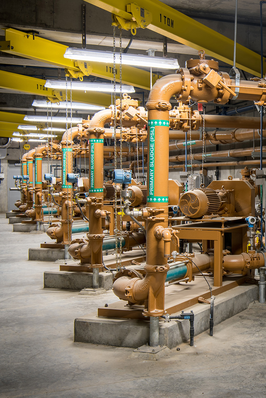 Six rows of orange pipes labeled “primary sludge” run across a large concrete room. 