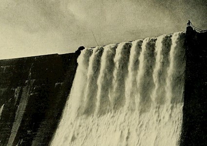 Picture shows a black and white image of water rushing over the edge of a dam. 
