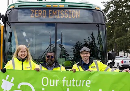 three people smile in front of a bus with a sign promoting electric buses