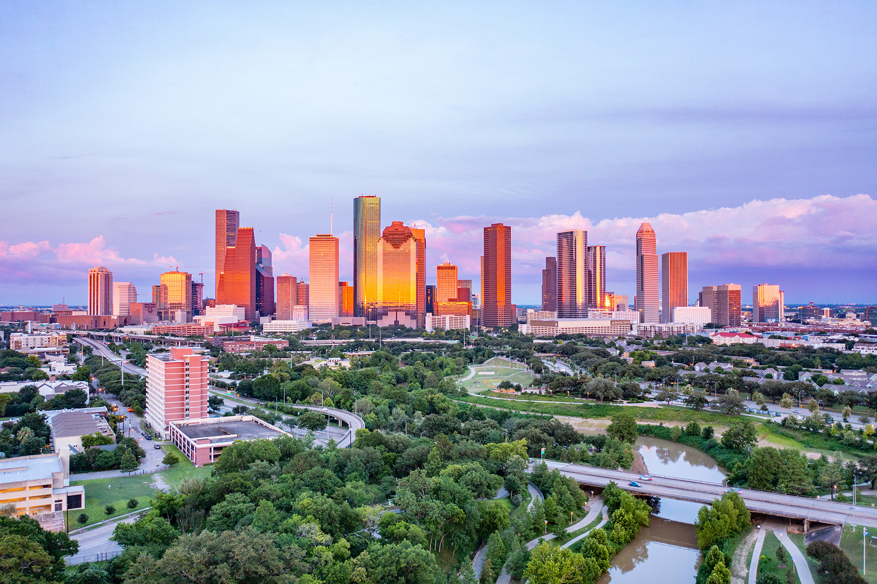 In the foreground, a park with trees and a highway above it. In the background, skyscrapers stand amidst a partly cloudy sky.