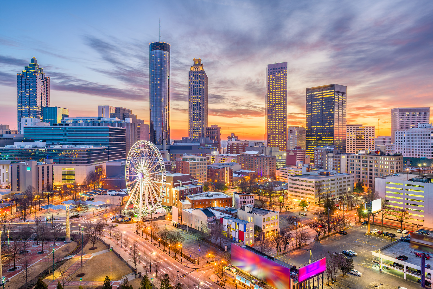 An aerial view of Atlanta’s towering skyscrapers and shorter buildings make up blocks of the city’s downtown. In the foreground are a highway, Ferris wheel, and a parklike setting with paths, trees, and sculptures.