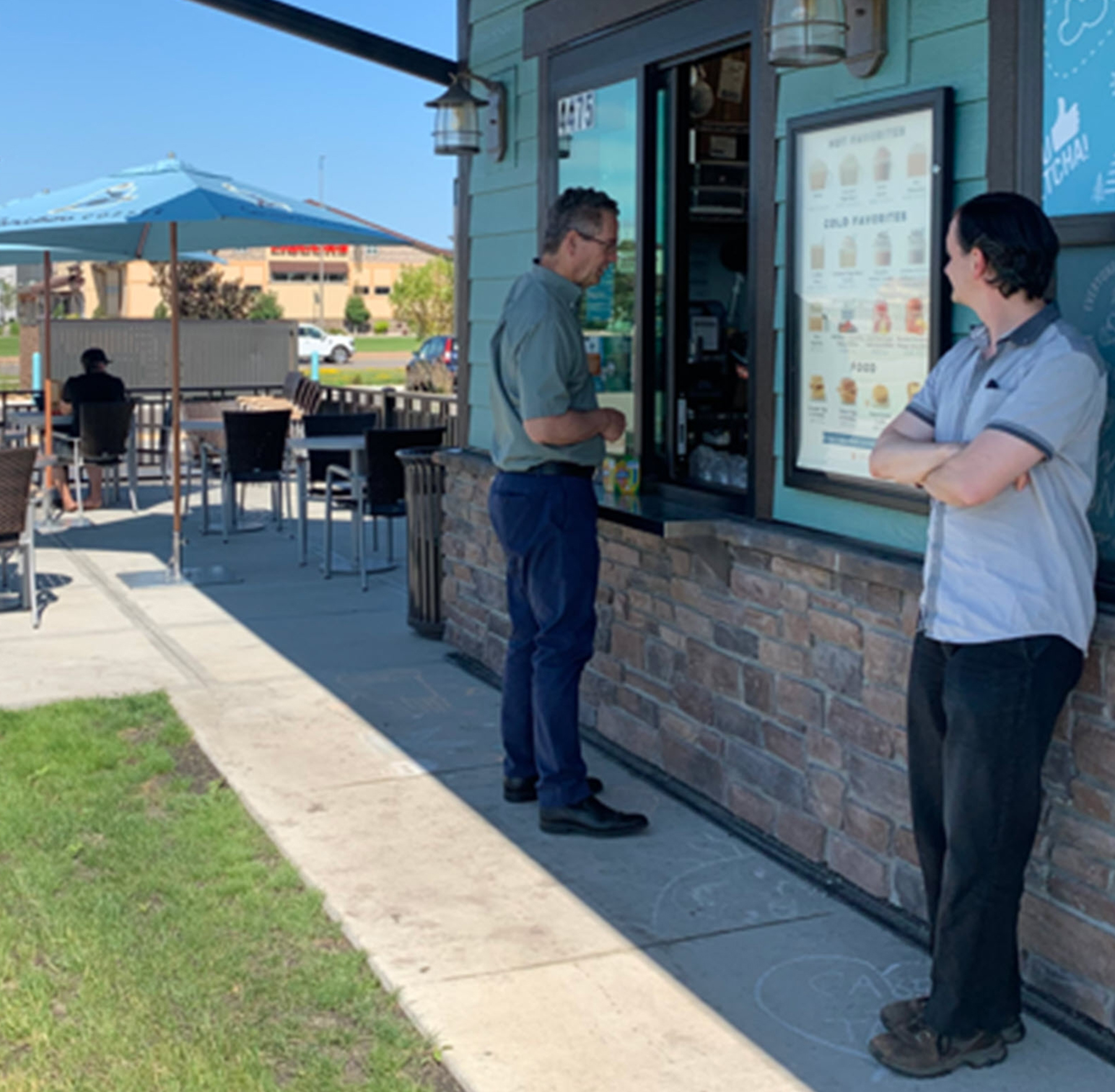 two men appear at a walk-up window with another sitting at an outdoor table in the background two men appear at a walk-up window with another sitting at an outdoor table in the background