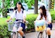 Two women ride bicycles with white baskets down a bike path in Japan. A line of green shrubs separates them from parked cars and traffic.
