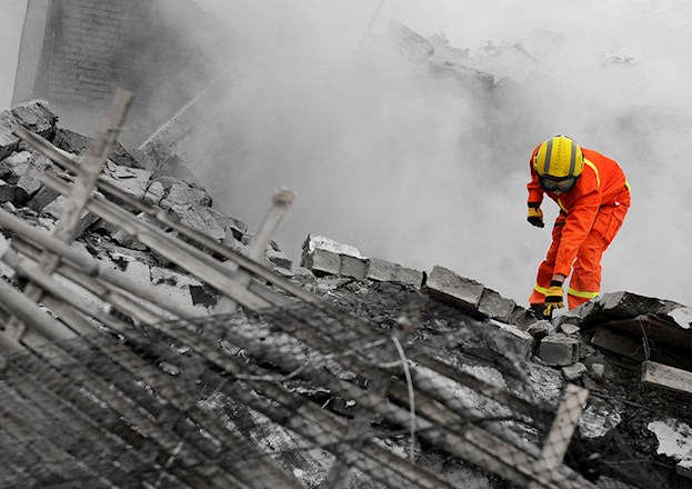 A first responder in full orange personal protective gear and thick goggles stands on a mound of collapsed structure.
