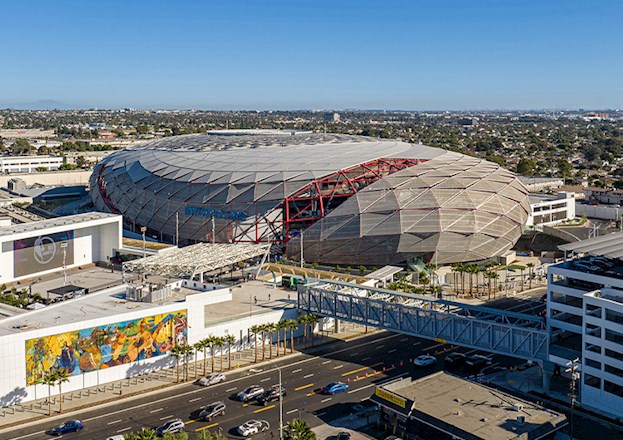A sports arena sits amid the cityscape. There are walkways to the arena and many open entertainment and gathering spaces in front of the stadium.