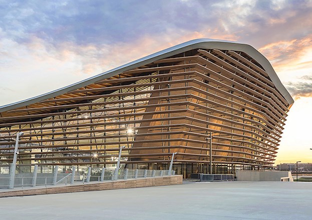 The finished building displays the timber elements comprising the sides and roof. A white pedestrian bridge is seen in the foreground and surrounding buildings in the background.