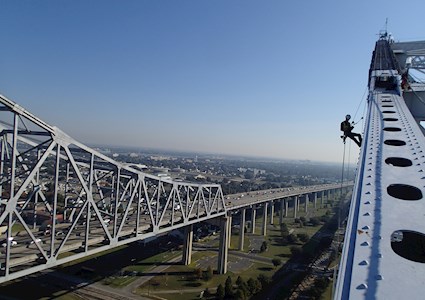In the foreground, a person in full safety gear uses a rope system to inspect the top of a bridge. In the background is a multi-lane highway.