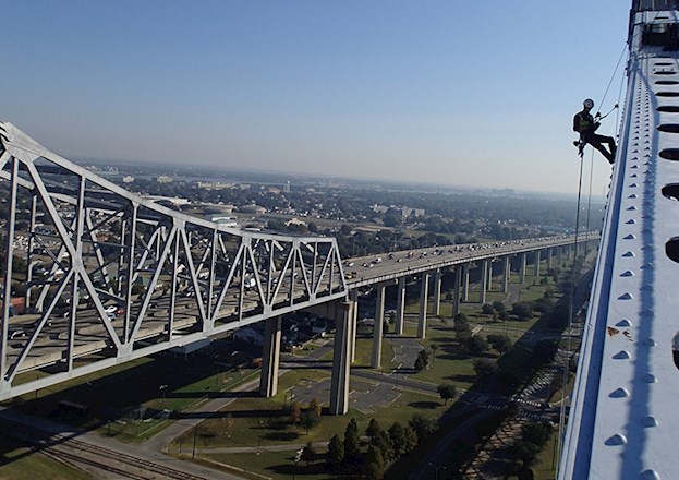 In the foreground, a person in full safety gear uses a rope system to inspect the top of a bridge. In the background is a multi-lane highway.