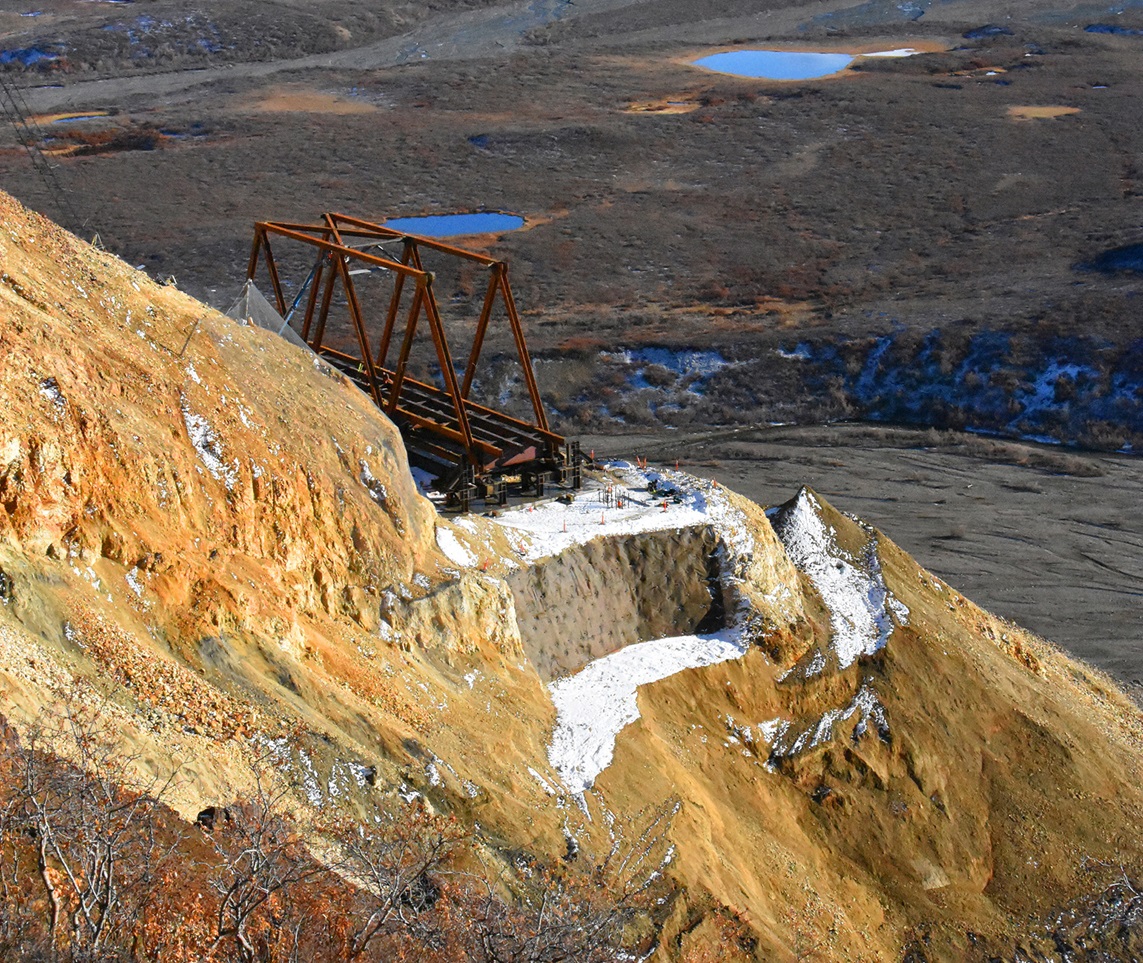 Photograph shows a steel truss bridge under construction on a mountainside. One side of the foundation can be seen. 
