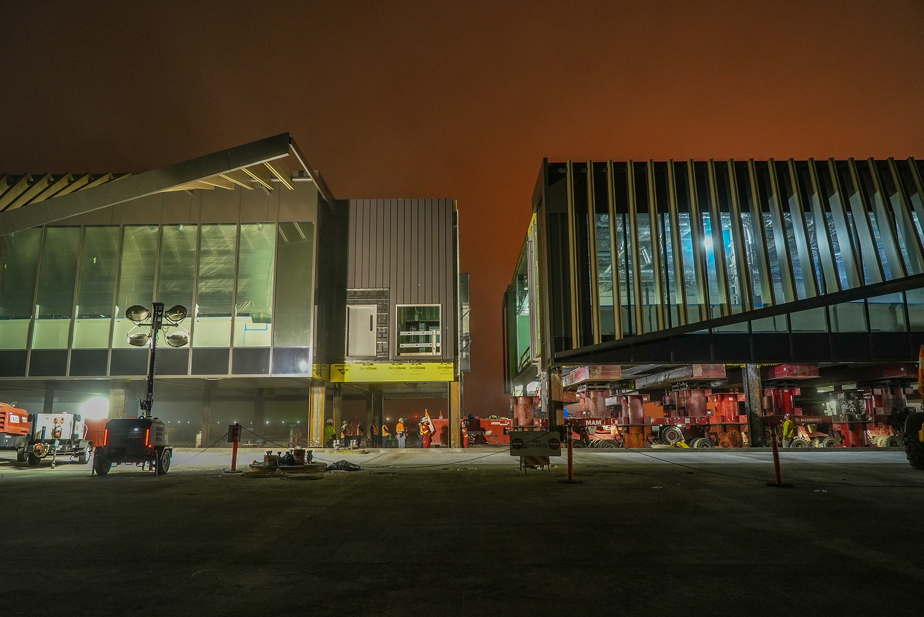 Two segments are placed side by side to be connected. It’s nighttime and there are large flood lights illuminating workers and equipment. 