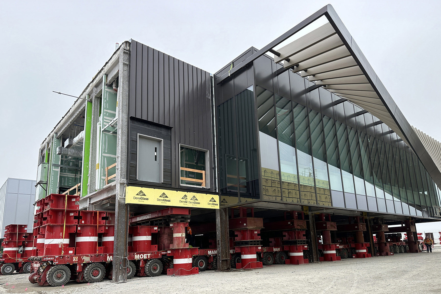 Red and white self-propelled modular transporters lift a segment for transport. You can see the glass facade and angled shades. 
