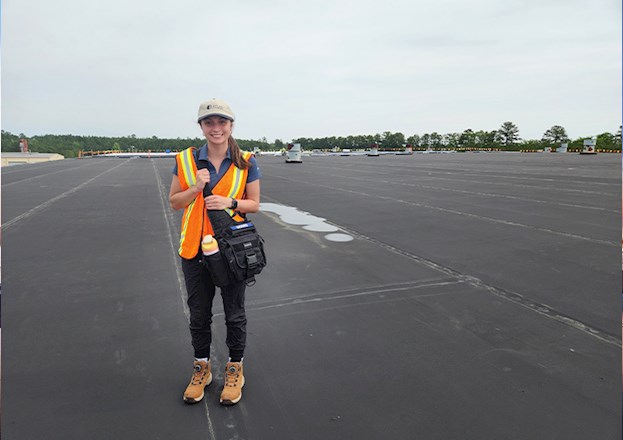A smiling young woman, in safety vest and Atlas cap, stands atop the roof of a building.