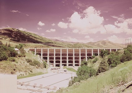 Photograph shows a multiple-arch dam set amidst trees and hills.