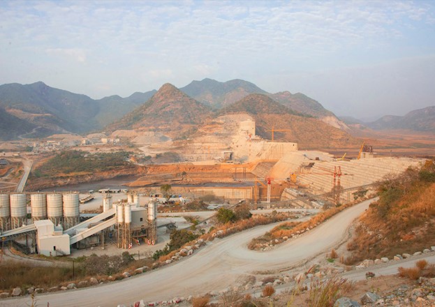 The photograph shows the massive dam under construction in an arid landscape.