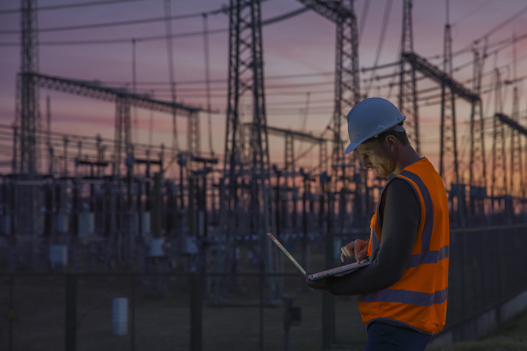 Man checks his computer while standing among power transmission lines.