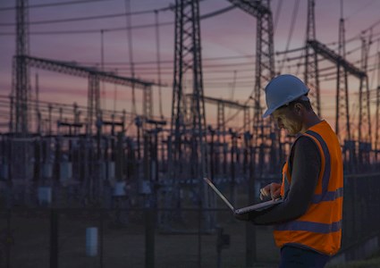 Man checks his computer while standing among power transmission lines.
