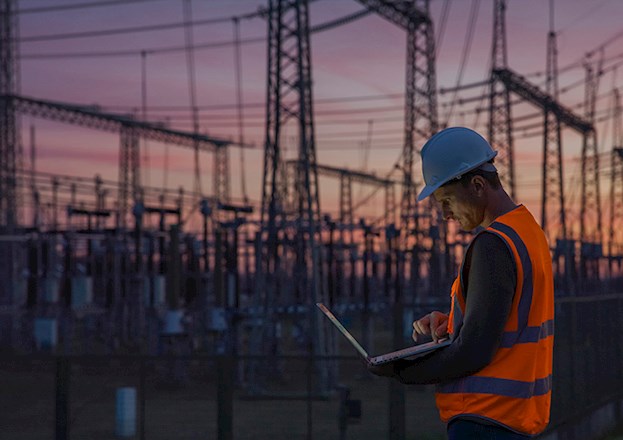 Man checks his computer while standing among power transmission lines.