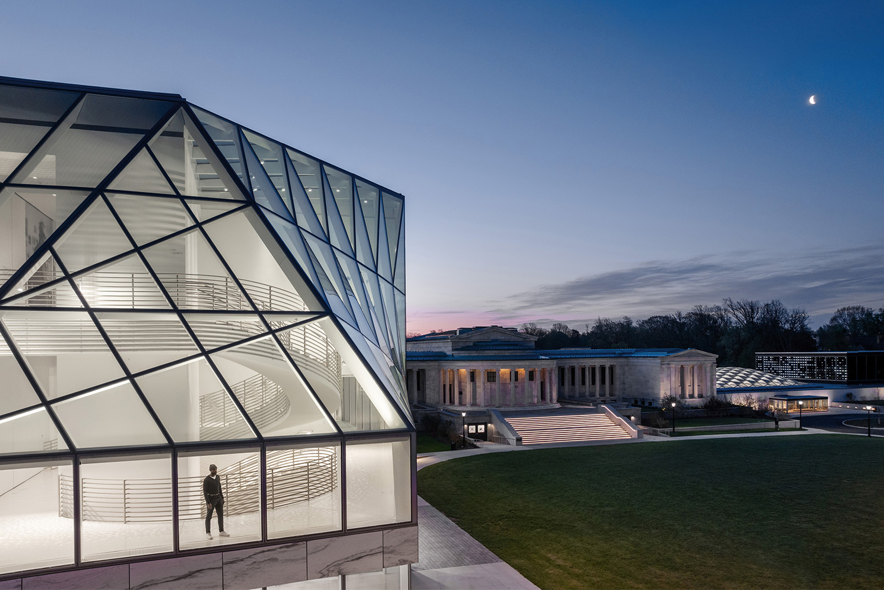 A man stands inside a glazed building, looking out at night. Behind him, a large spiral staircase rises through the building’s floors.