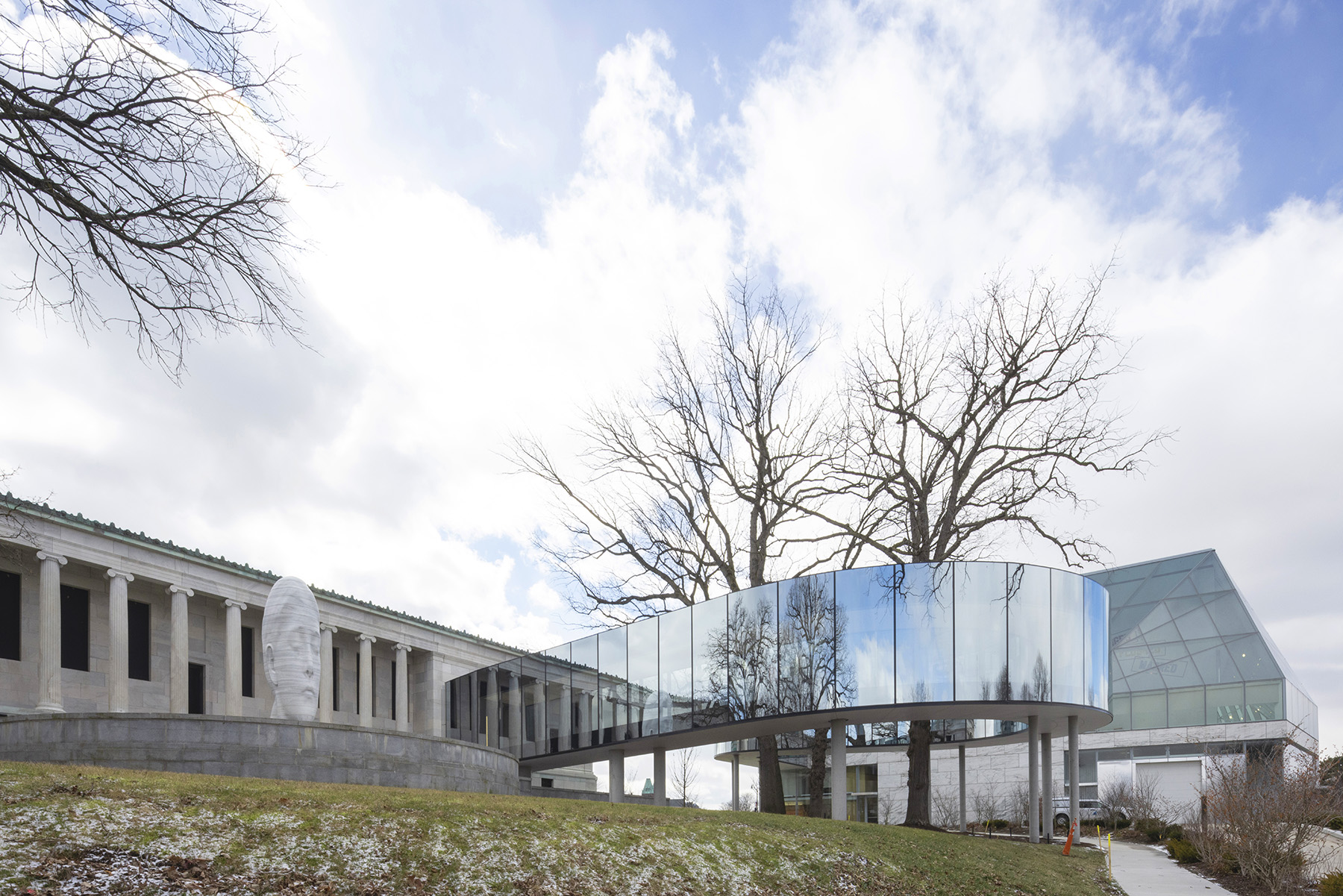 An enclosed, glazed bridge extends from one building and wraps around several tall trees before connecting to another building.