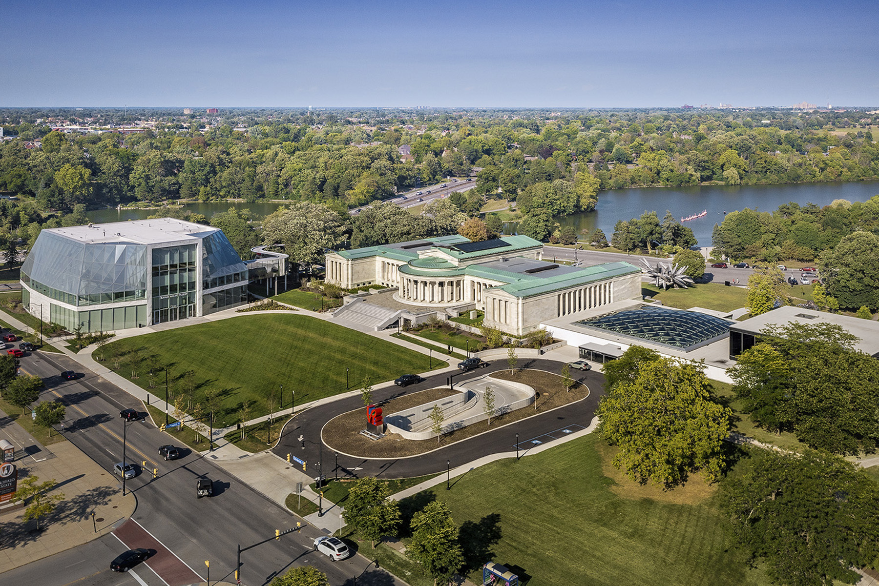 An aerial photograph shows the Buffalo AKG Art Museum campus, including the modern-looking glazed new building, the columned facades of the 1905 building, and the glazed canopy covering the now-enclosed courtyard. The new green lawn covering the underground parking garage is shown in the foreground and a body of water is visible in the background.