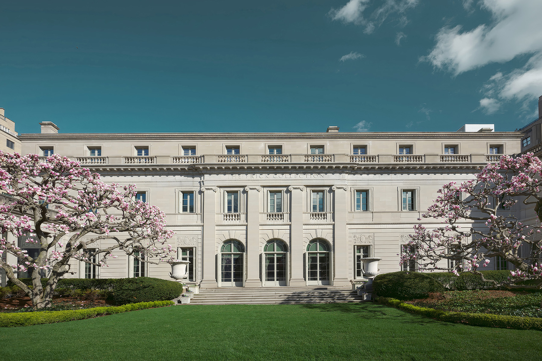The stone facade of a multistory mansion is shown. There are flowers on the trees and steps leading down to beautiful green grass.