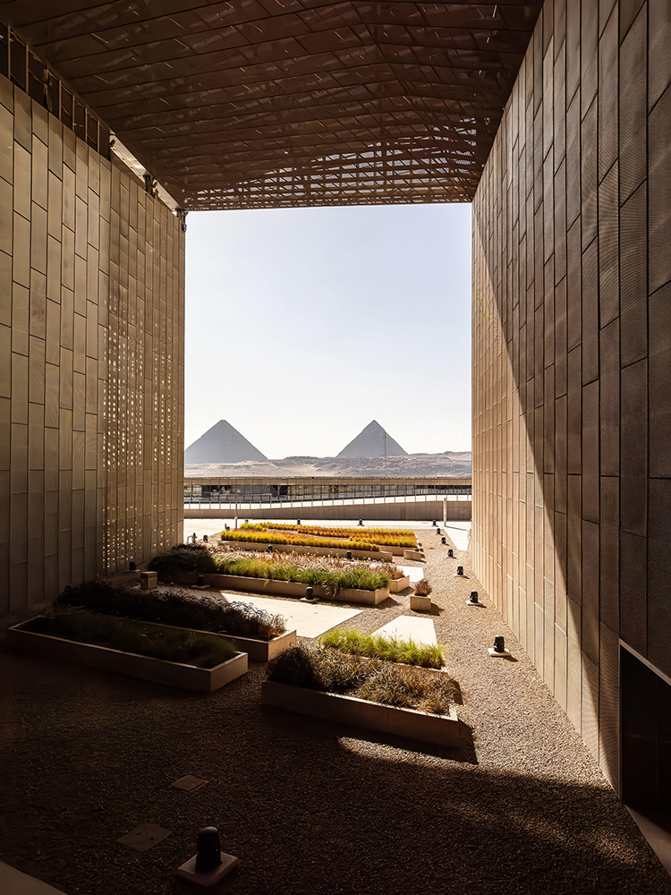 In the distance, two pyramids can be seen. In the foreground, there are flower beds arranged on a diagonal. A steel diagrid roof can be seen above the plantings. 