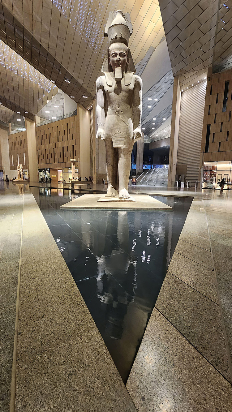 A tall concrete statute of a pharaoh stands on a plinth inside a museum. 
