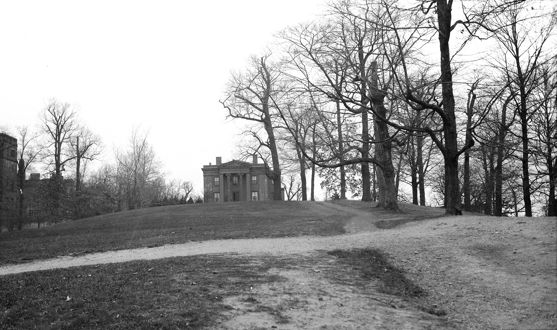 A black-and-white image shows a large grassy lawn in the foreground with a path that leads up to a single two-story Gothic style building. Trees line both sides of the lawn.