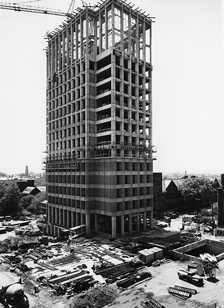 A black-and-white image shows a multi-story building under construction. Cranes and scaffolding sit at the top of the building. 