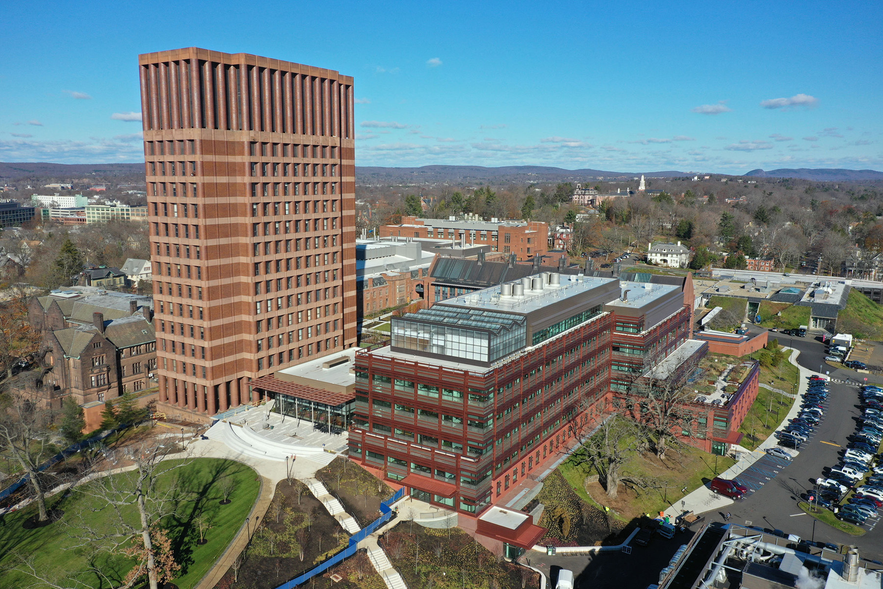 An aerial view of the entire Science Hill complex shows several buildings of differing heights, surrounded by grassy areas and parking lots with cars. In the background, trees and other buildings.