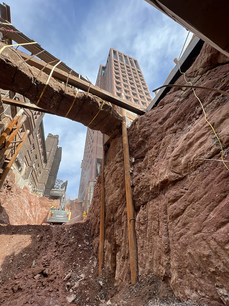 A photo taken from underground of bedrock excavation. Construction workers and two diggers clear dirt from the site.  