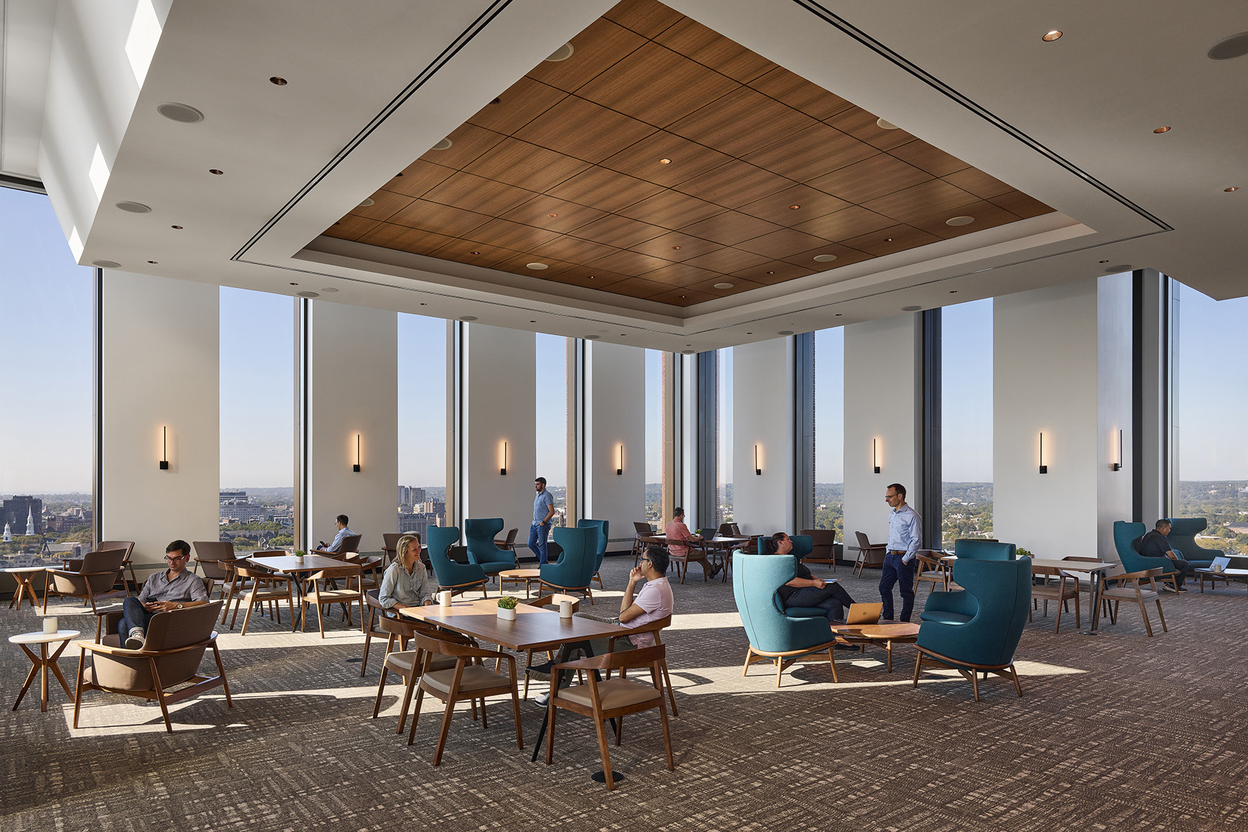 A photograph of a room with mid-century modern style, warm-wood-toned tables and chairs spaced out around the room. In the background, floor-to-ceiling windows alternate with floor-to-ceiling walls, each with a single modern-style light fixture.