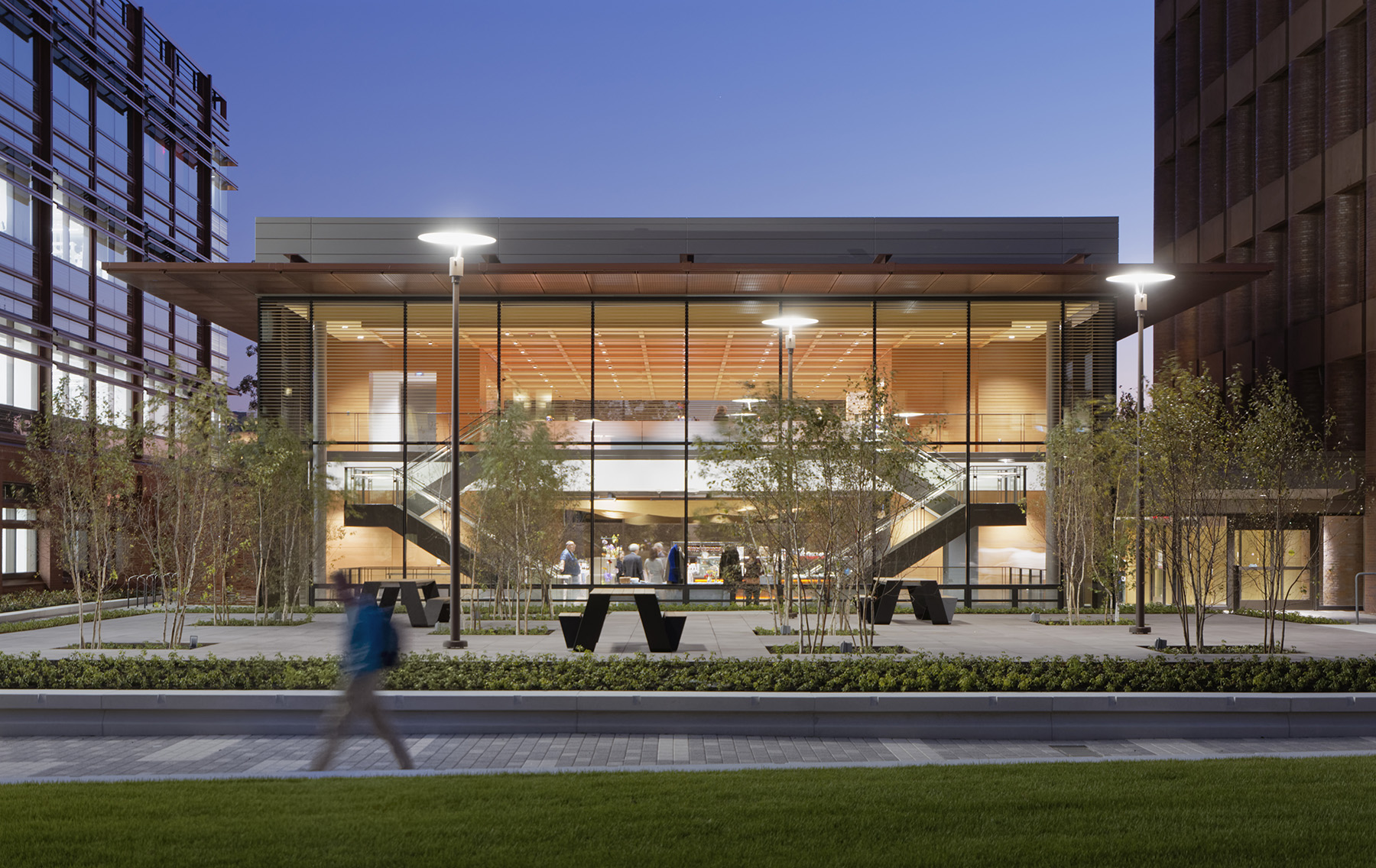 A two-story building with floor to ceiling glass windows has two parallel staircases. Outside the building, there is a stone patio with trees and seating. A person walks in the foreground.
