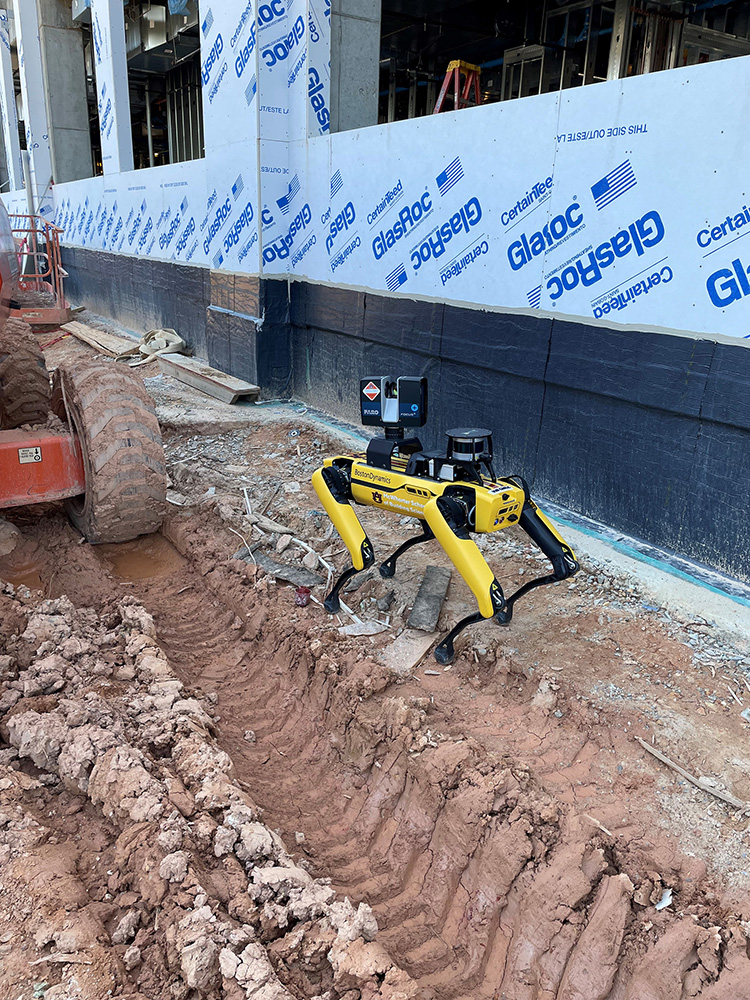 A yellow and black four-legged robot has cameras and sensors on its back as it walks through a construction site. 