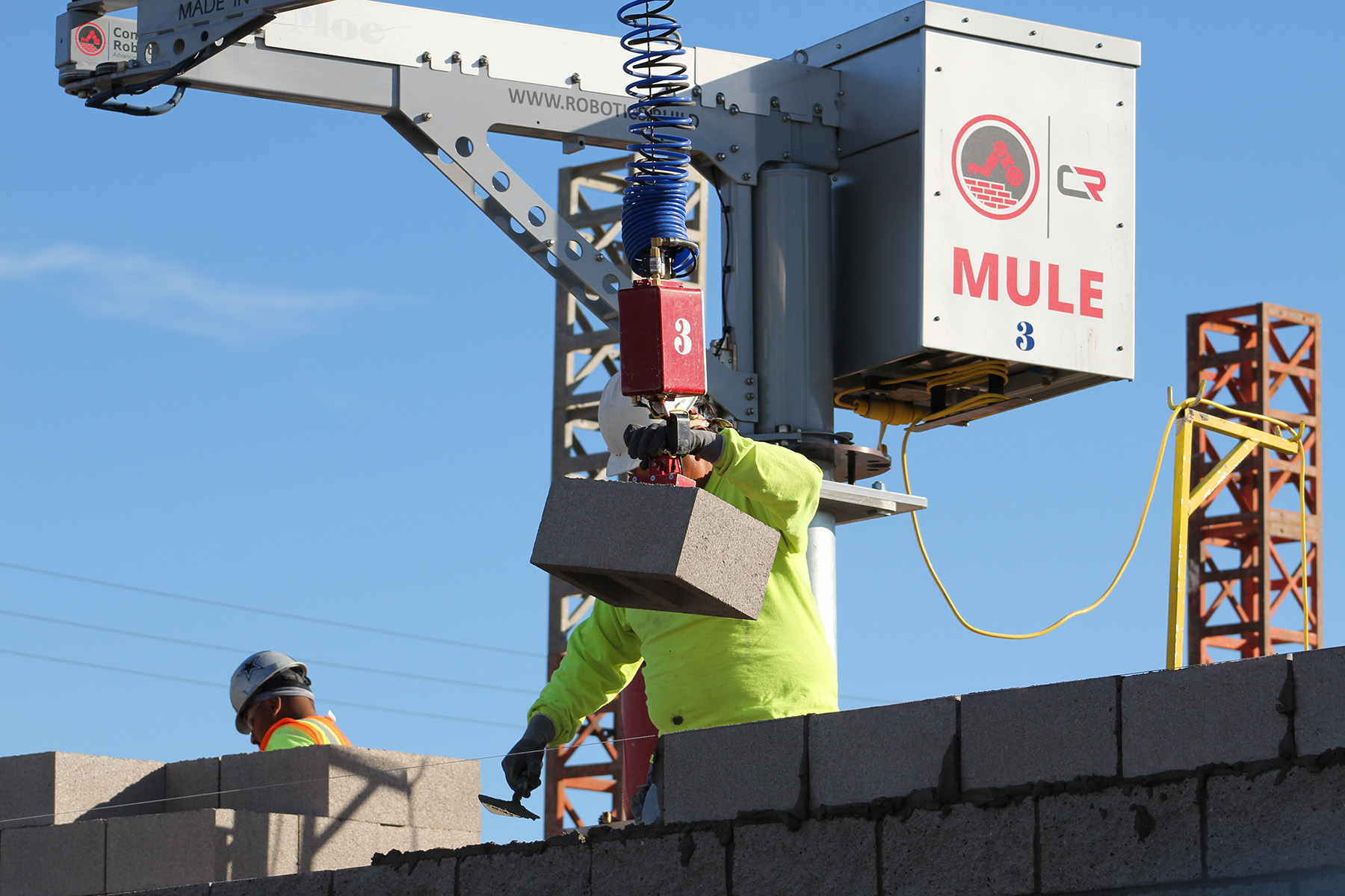 A construction worker uses a machine with the word “MULE” in red lettering to lift concrete bricks. The worker is building a wall. 