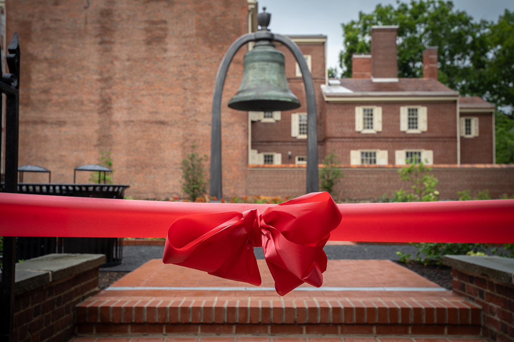 A blurred image of a six-ton bronze bell that hangs in a park, surrounded by brick buildings. A red bow is added over the image. 