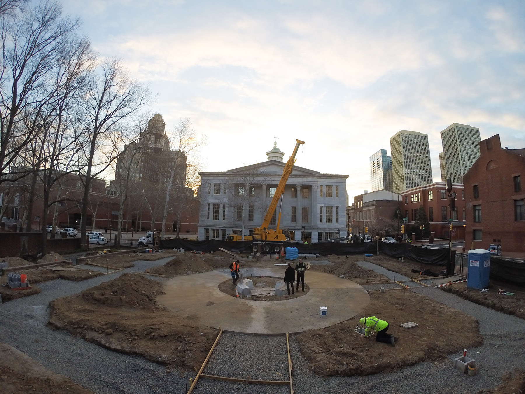 Three men stand in the middle of a concrete circle. There are dirt piles surrounding them and a large yellow crane behind them. Behind the crane are both skyscrapers and historic brick buildings. 