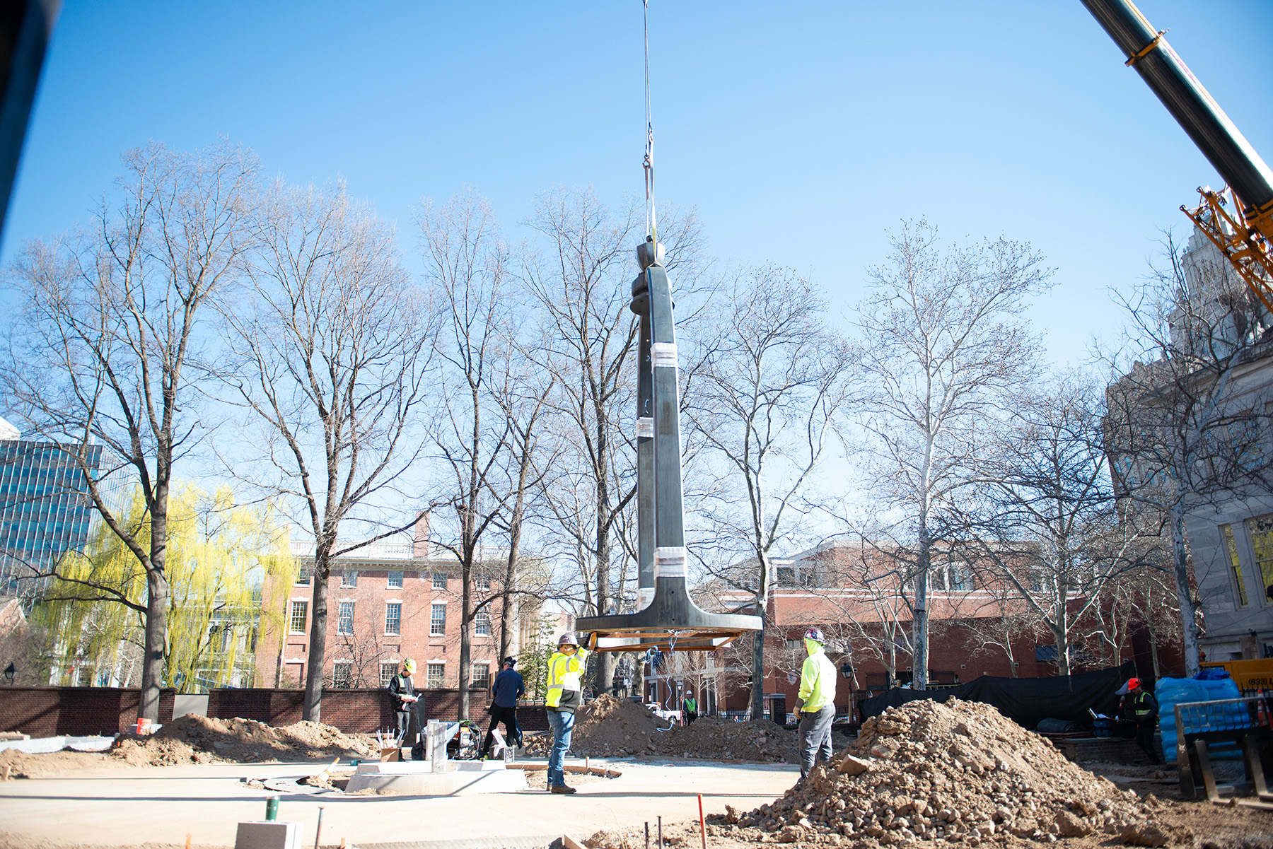 A crane holds the iron arch a few feet above the ground. Four men work on the site in the background. 